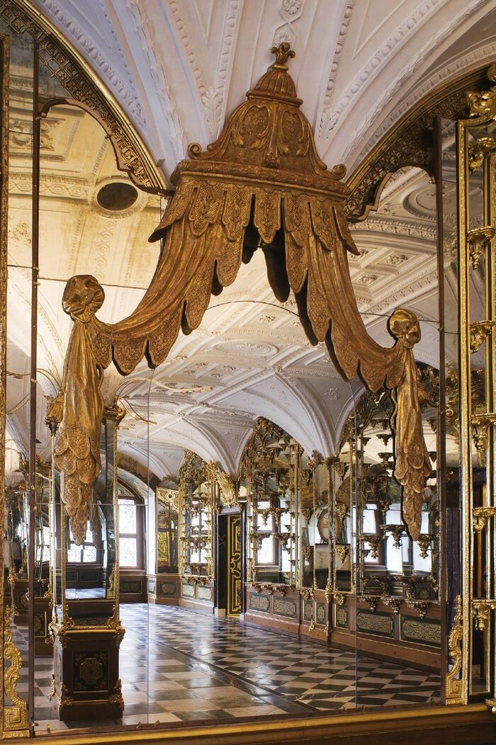 In einem Spiegel mit Goldbaldachin spiegelt sich ein Saal mit Säulen, gewölbter Stuckdecke und verspiegelten Wänden mit Konsolen auf denen Objekte stehen