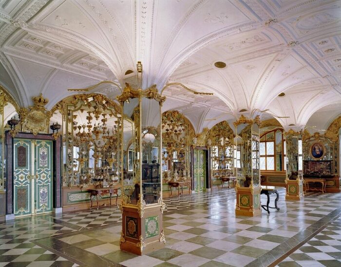 Saal mit Säulen, gewölbter Stuckdecke und verspiegelten Wänden mit Konsolen auf denen Objekte stehen