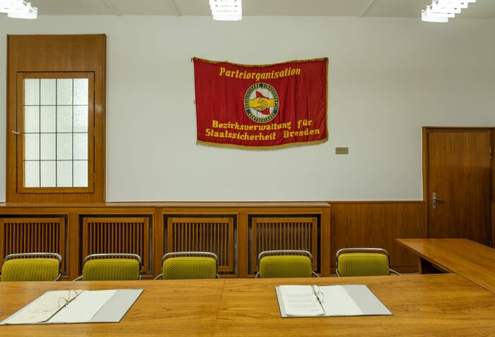 Versammlungszimmer mit Beratungstisch und Stühlen. Dahinter ein Banner mit der Aufschrift Parteiorganisation Bezirksverwaltung der Staatssicherheit Dresden