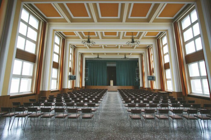 Festsaal der Stasi mit Blick über die Stuhlreihen auf die Bühne. Der Saal hat hohe Fenster. Die ist mit farbigen Kassetten geschmückt.