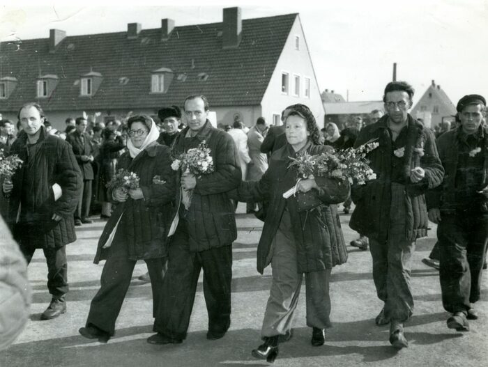 Mara Jakisch und weitere Personen bei ihrer Ankunft in Friedland 1955. Sie tragen Blumen im Arm und der Weg ist von winkenden Menschen gesäumt. Im Hintergrund niedrige Häuser