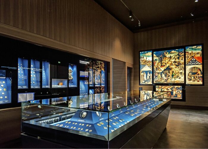 Blick in die Ausstellung, Vitrinen und Glaskästen mit Münzen. An einer Wand ein mehrteiliges, beleuchtetes Glasfenster mit bäuerlichen Szenen