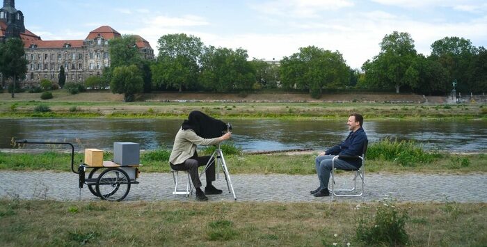 Am Elbufer sitzen zwei Männer. Der eine fotografiert den anderen mit einer alten Standkamera. Der Kopf des Fotografen steckt unter einer Decke. Auf der anderen Flussseite die Staatskanzlei in Dresden