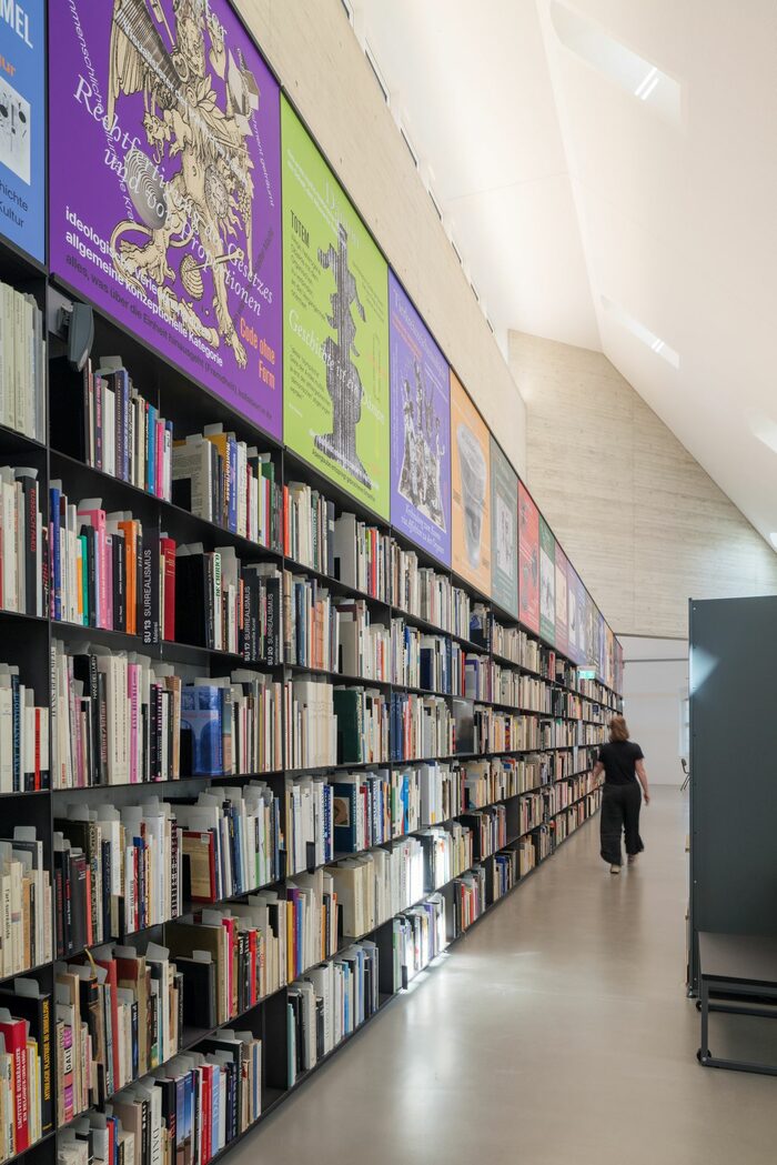 In einem langen Gang erstreckt sich ein Bücherregal, hoch voll mit Büchern, bis ans andere Ende des Raumes