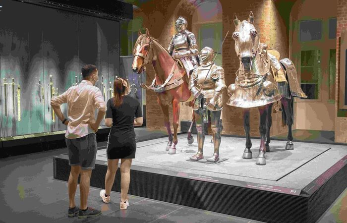 Ein Pärchen betrachtet Ausstellungsstücke auf einem Podest. Zu sehen sind zwei Holzfperde und zwei Ritter, die alle mit silberglänzenden Harnischen bekleidet sind.