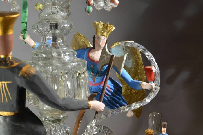 Bildausschnitt eines Glas-Kronleuchters. Im Bild hängt wie schwebend die Glasfigur einer Frau mit Krone
