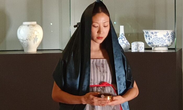 Vor Vitrinen mit Porzellan steht eine asiatische Frau. Sie trägt ein Tuch auf dem Kopf und hält in der Hand ein kleines Gefäß.