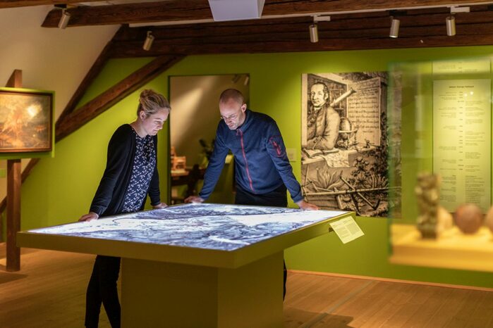 Ein Ausstellungsraum in einem alten Haus mit Holzbalken. Zwei Besucher betrachten den interaktiven Teil der Ausstellung.