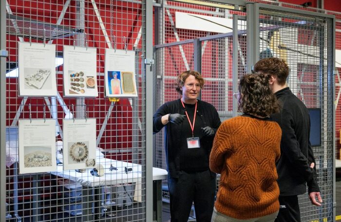 Im Raum steht ein mit Metallgittern abgetrennter Bereich. An den Gittern hängen Skizzen und eine Frau mit Arbeitshandschuhen ist im Gespräch mit zwei weiteren Personen.