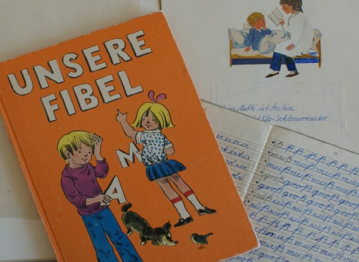 Eine orangefarbene Schulfibel mit zwei Kindern, einem Hund und einem Vogel auf dem Deckel, dazu eine Heftseite aus einem alten Schulheft, gefüllt mit Schreibübungen