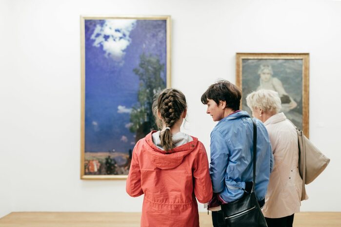 Menschen stehen in einem Ausstellungsraum mit Gemälden. Ein hochformatiges Bild zeigt einen Baum und weitere Landschaftsdetails vor einem hohen Himmel. .