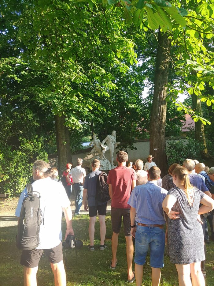 Im sommerlichen Park gehen bunt gekleidete Menschen auf eine Skulpur mit zwei sagenhaften Figuren zu.